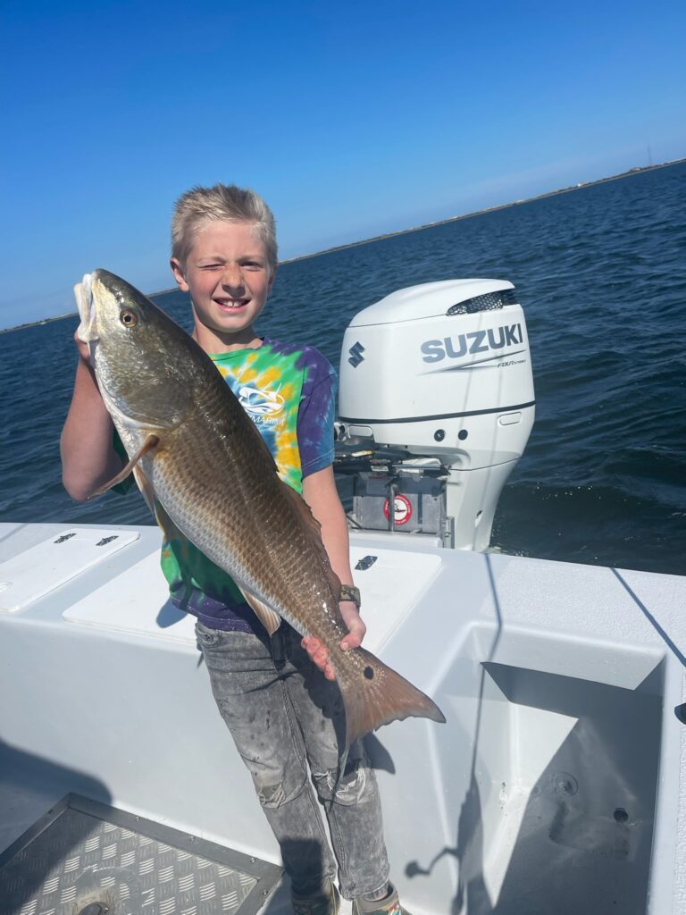 A young boy proudly displays a fish he caught on a fishing charter boat trip. He wears a life jacket and holds the fish with both hands, smiling widely. In the background, blue water and other boats can be seen. It's an excellent family activity for kids to learn about fishing and enjoy the outdoors.